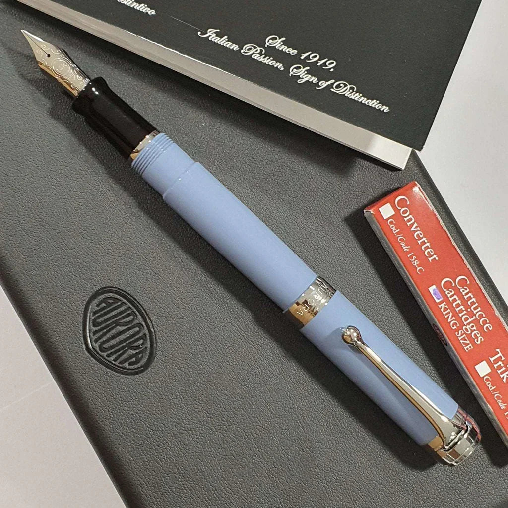 Light blue fountain pen with silver trim on a black notebook, converter and cartridge box nearby.