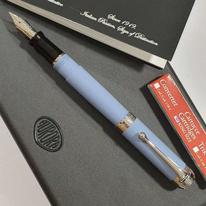 Light blue fountain pen with silver trim on a black notebook, converter and cartridge box nearby.